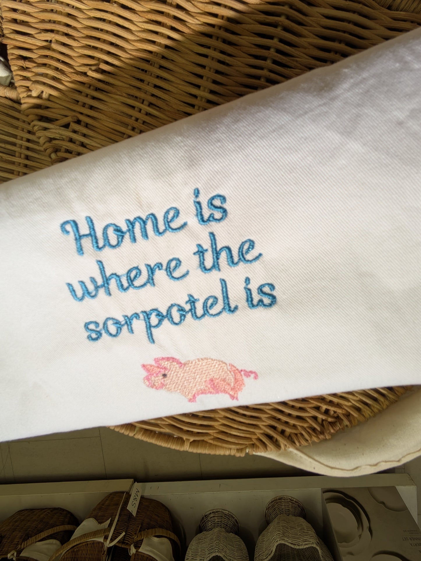 Embroidered towel with 'Home is where the sorpotel is' text and a pig illustration on a woven basket background.